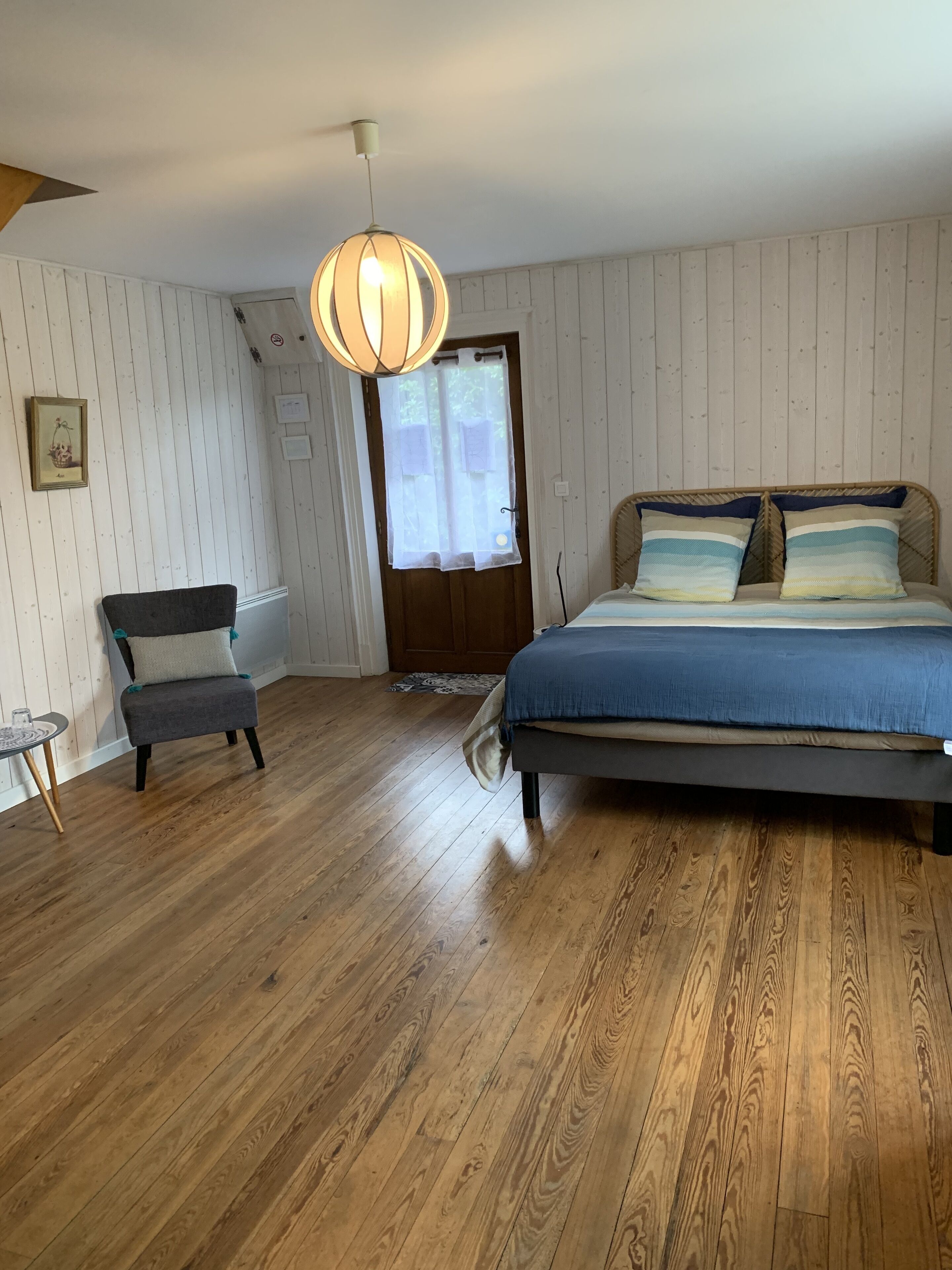 Un dormitorio con piso de madera, una cama con ropa de cama azul, una silla, una lámpara colgante y una puerta con ventanal.
