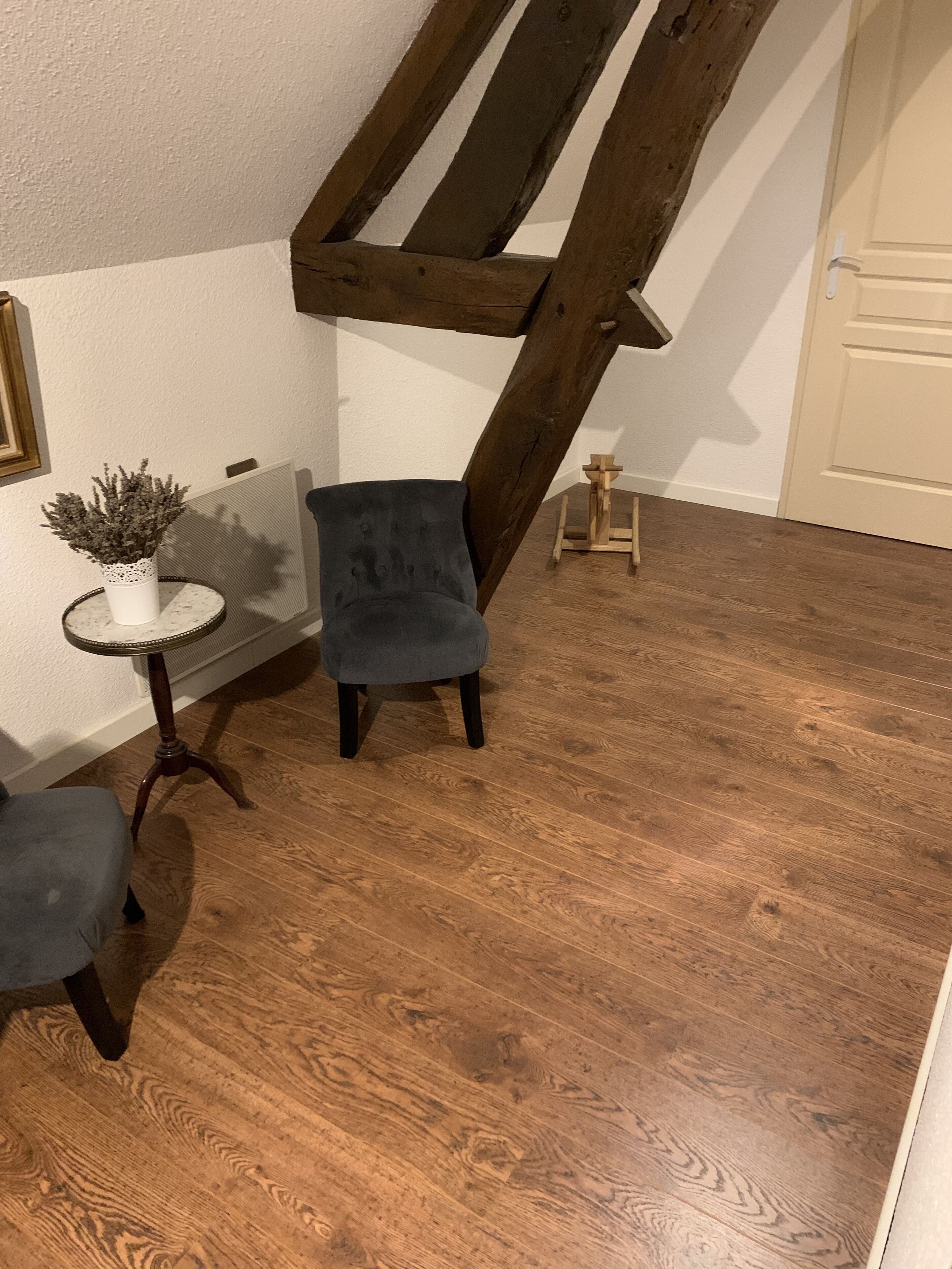 A room with a wooden floor, a grey upholstered chair, a small white table with a plant, and a white door.