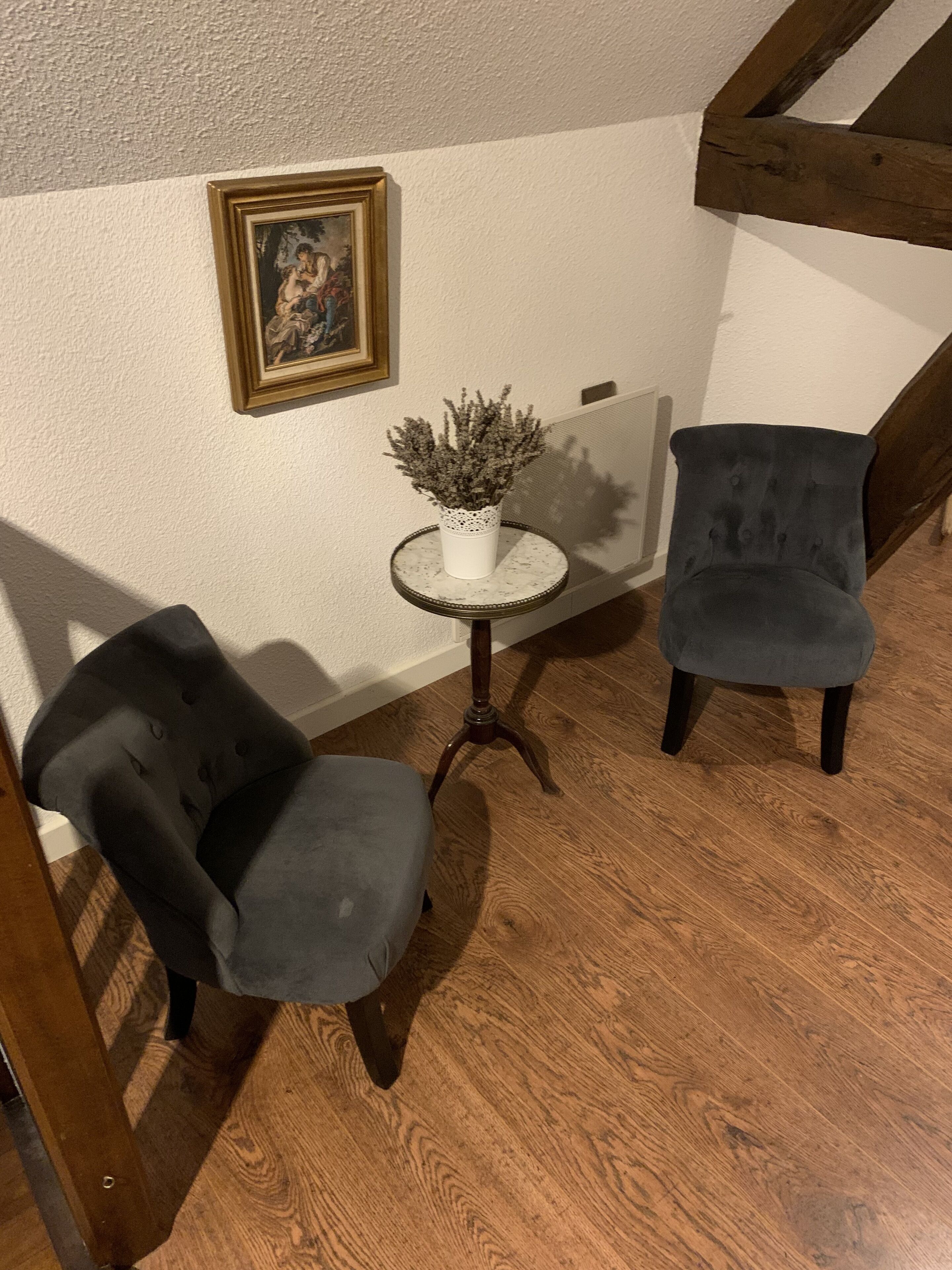 A corner room with two dark grey upholstered chairs, a small round table with a white vase of dried flowers, and a framed picture on the wall.