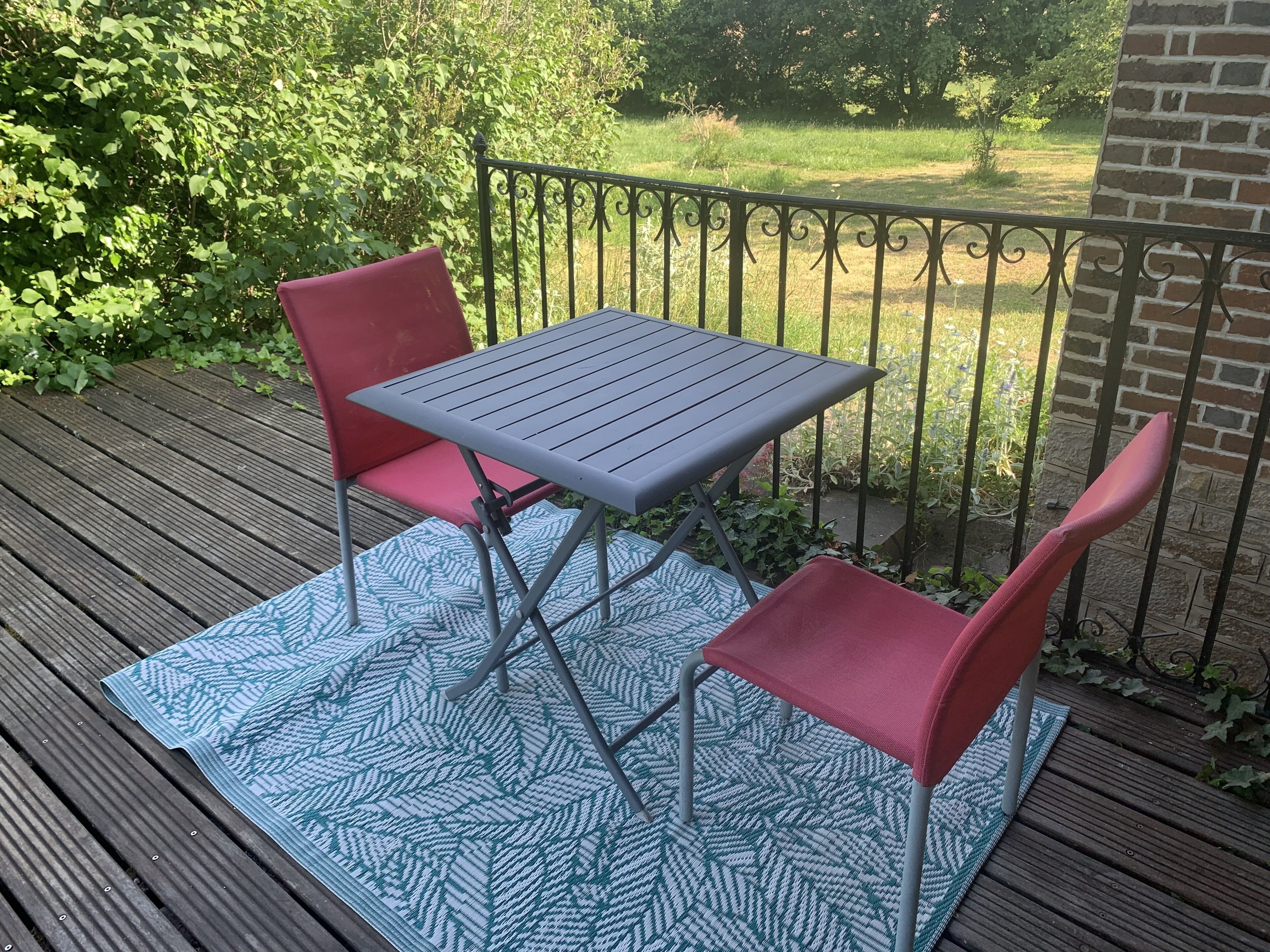 A deck with a metal table and two red chairs, a patterned rug, and a brick wall in the background.