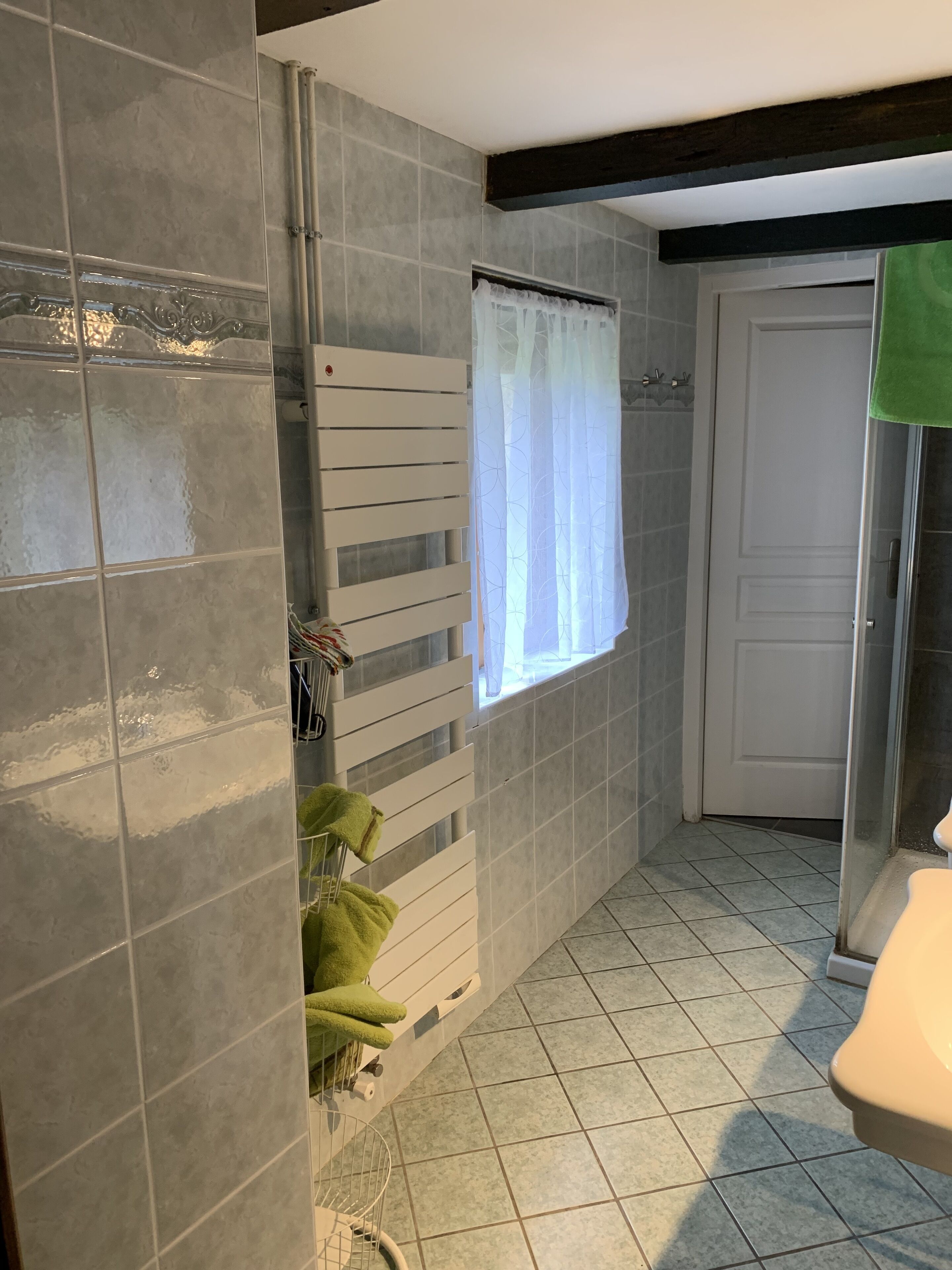 Une salle de bain avec des murs recouverts de carreaux gris, un porte-serviettes blanc, une fenêtre avec un rideau léger et une serviette verte sur le sol.