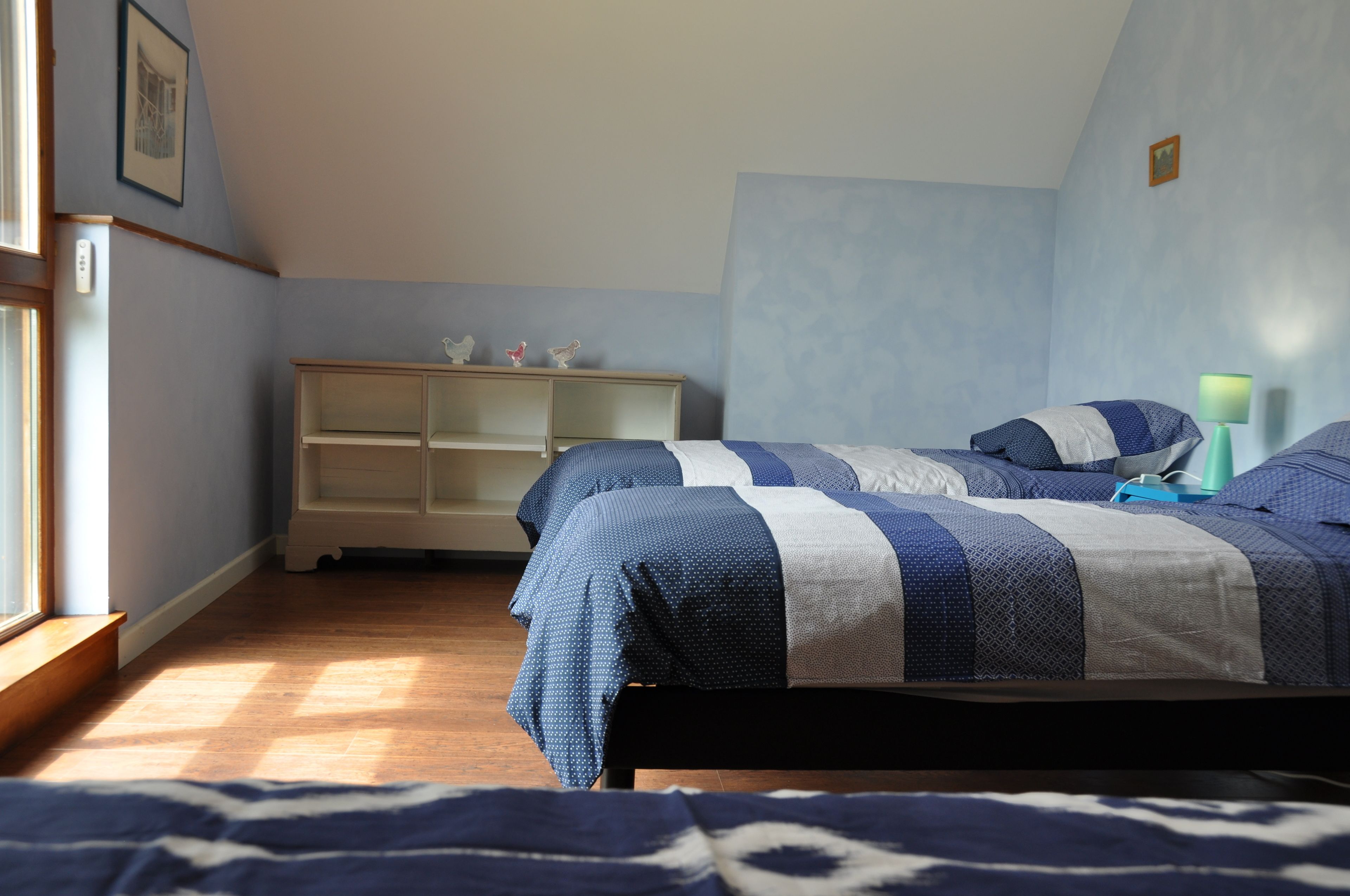 Une chambre avec deux lits, une commode en bois et une fenêtre laissant entrer la lumière naturelle.