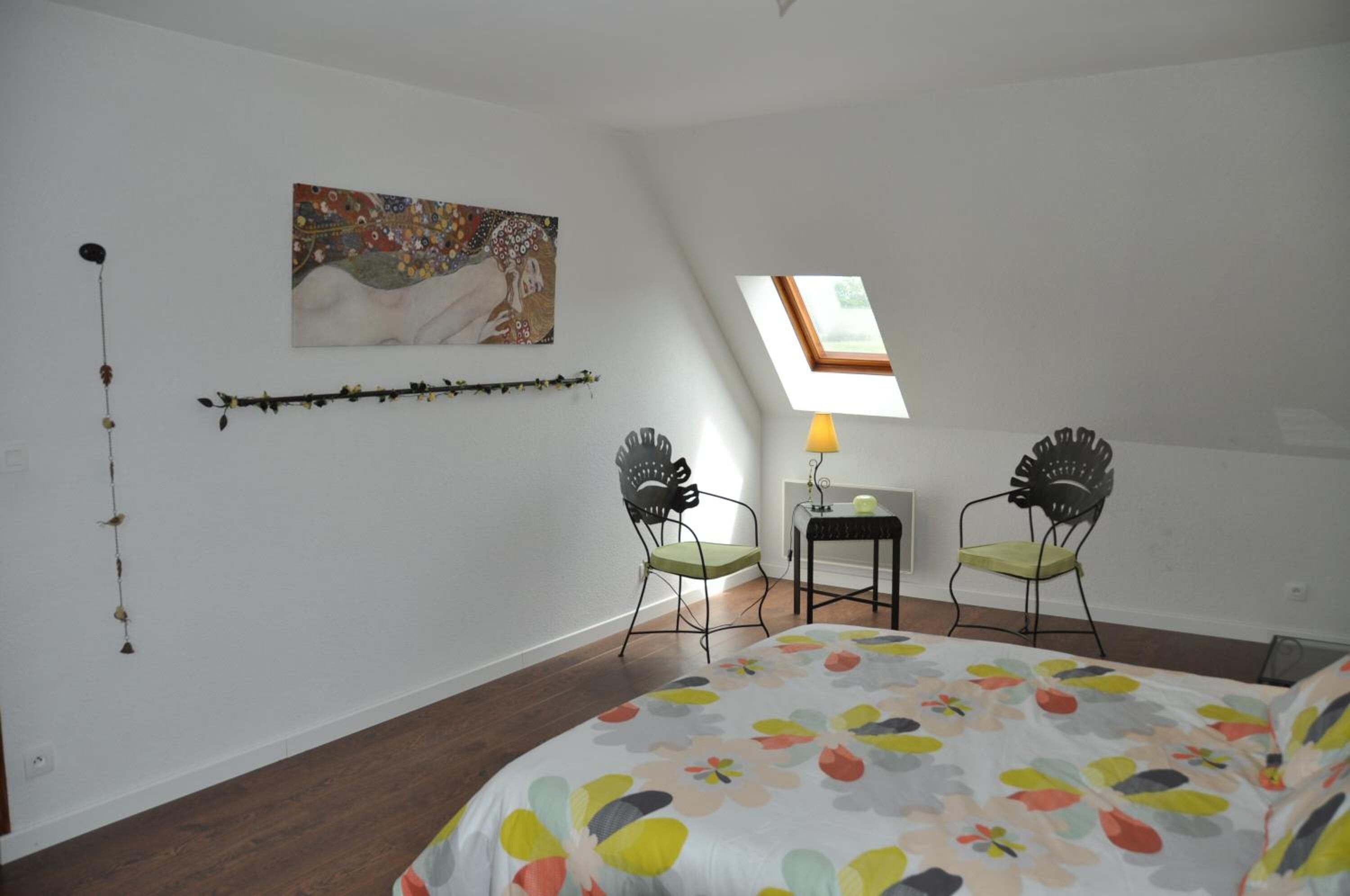 Une chambre à coucher comprenant un lit, deux chaises, une petite table, une lampe, un tableau et une lucarne.