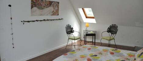Une chambre à coucher comprenant un lit, deux chaises, une petite table, une lampe, un tableau et une lucarne.
