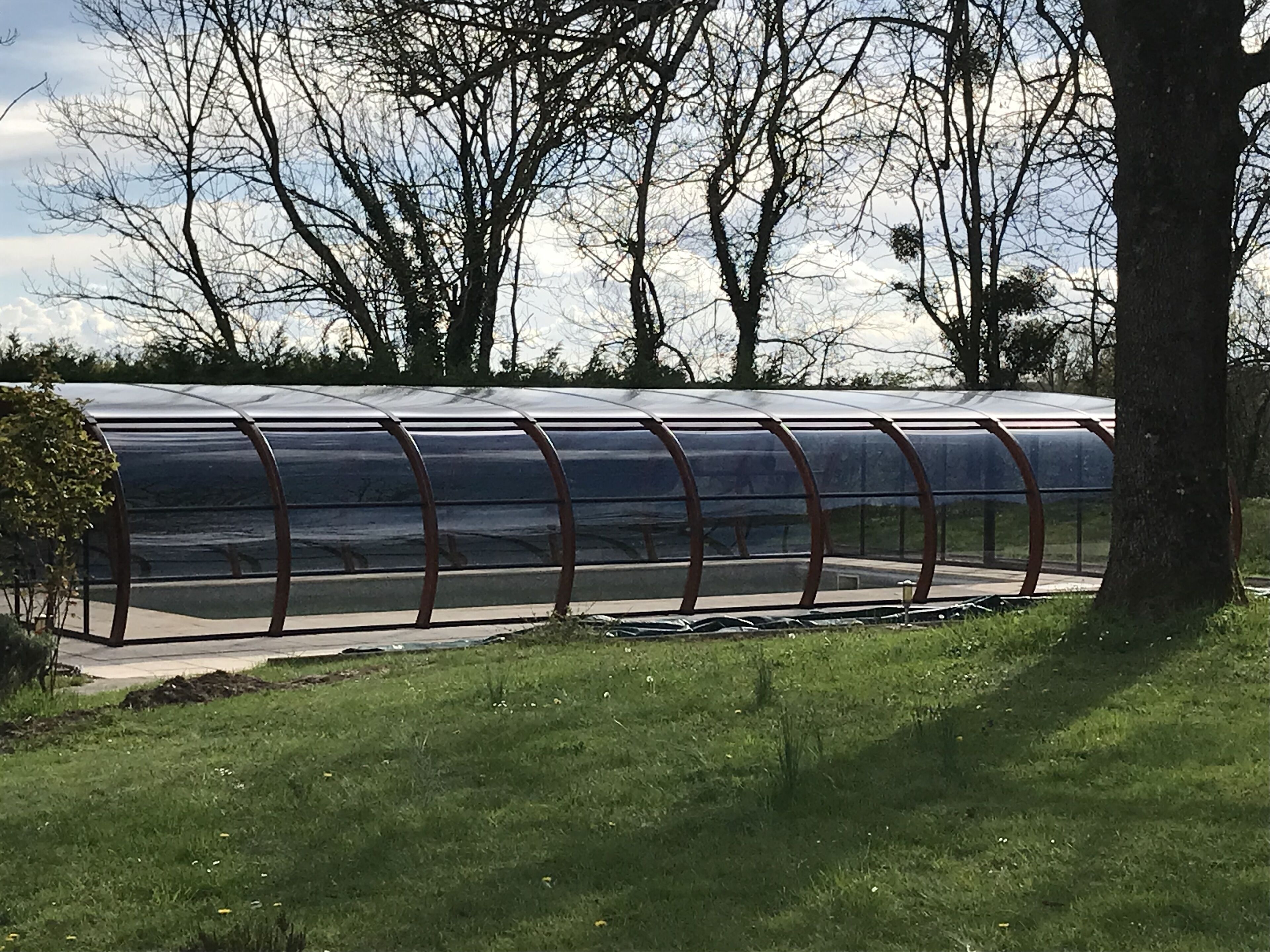 Une serre à structure transparente, dotée d’une armature métallique, entourée d’herbe et d’arbres.