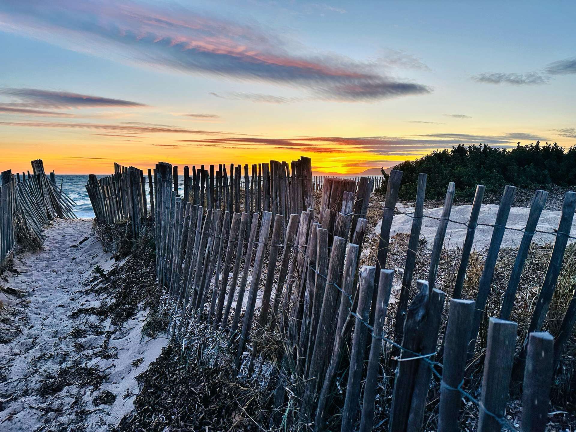 Ein hölzerner Zaun führt zu einem Strand, im Hintergrund ein Sonnenuntergang.