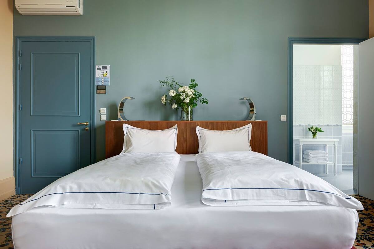 A double bed with white linens and a wooden headboard, a blue door, and a small table with a vase of flowers.