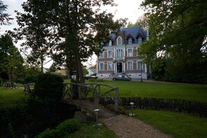Un grand bâtiment historique, doté d’une entrée imposante, entouré d’une pelouse bien entretenue et d’un pont en bois.