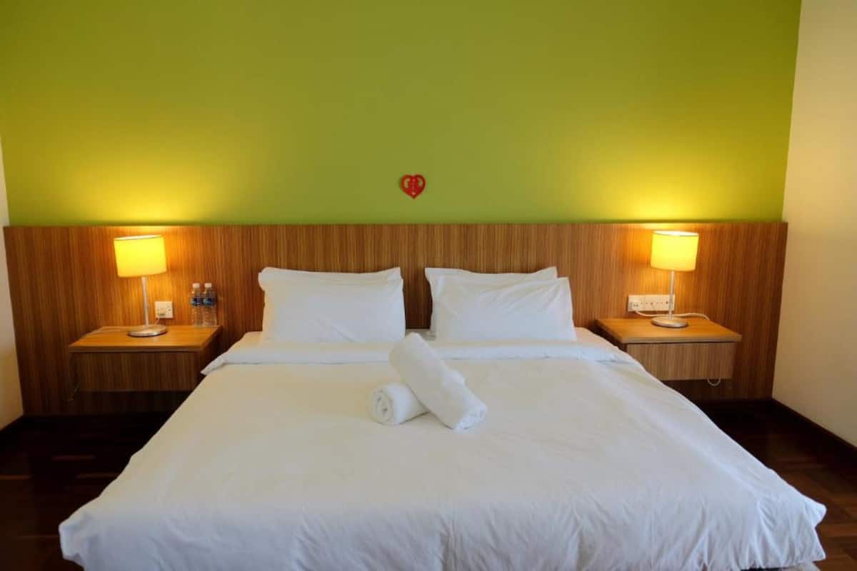 A hotel room with a wooden headboard, two bedside lamps, a heart-shaped decoration on the wall, and a neatly made bed with white linens.