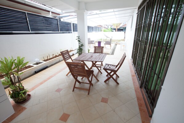 Un patio cubierto con mesa y sillas de madera, plantas en macetas y vista a la calle.