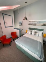 Un dormitorio con cama, dos sillas, un escritorio, un cuadro y una lámpara colgante.