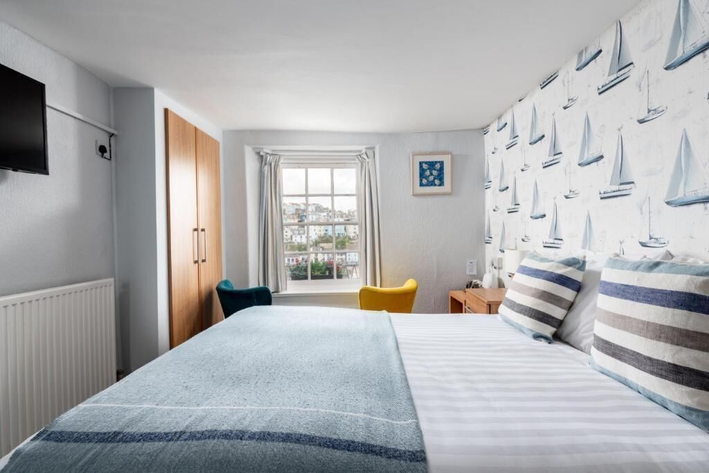 A hotel room with a bed, a window with curtains, a chair, a small table, and a wall-mounted TV.