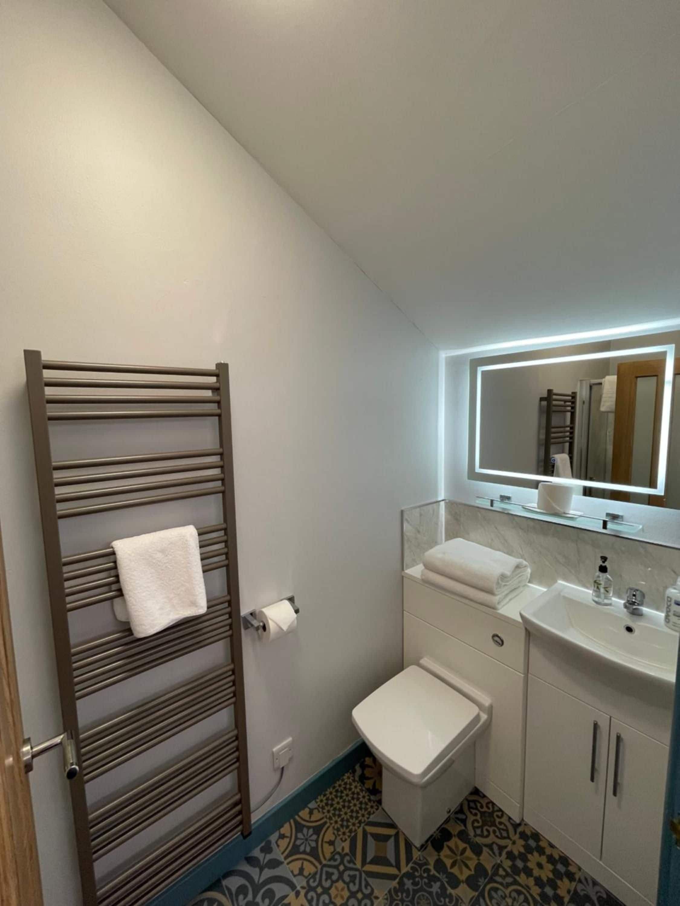 Un baño con toallero, inodoro, lavamanos y espejo con luz.