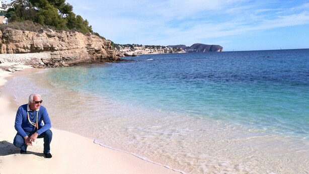 A person sitting on a sandy beach with clear water and rocky cliffs in the background. - APARTMENT NEW EXCEPTIONAL VIEW ON A COLINNE FACING THE SEA (a swimming pool) (benissa)