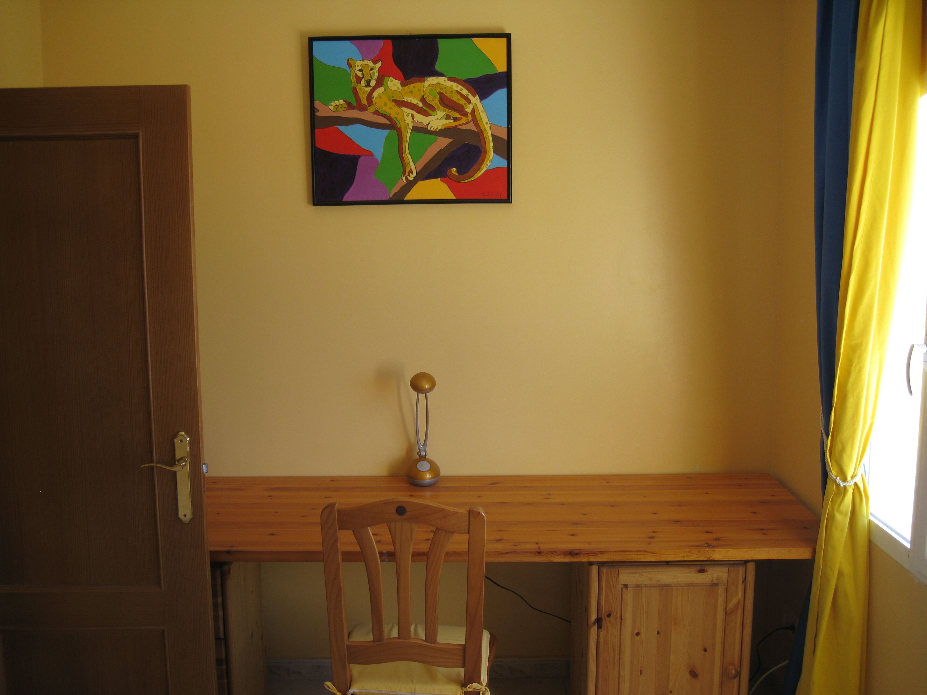 A room with a wooden desk, a chair, a door, and a colorful painting on the wall.