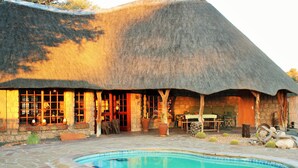 A thatched-roof building with a swimming pool, stone flooring, and outdoor seating. - Torgos Lodge (Koes)