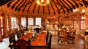 A rustic dining area with a long wooden table and chairs, stone walls, and a thatched roof. - Torgos Lodge (Koes)