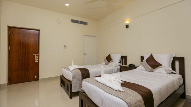 A hotel room with two beds, a ceiling fan, a door, and a light switch on the wall. - Le Noor (Kanayannur)