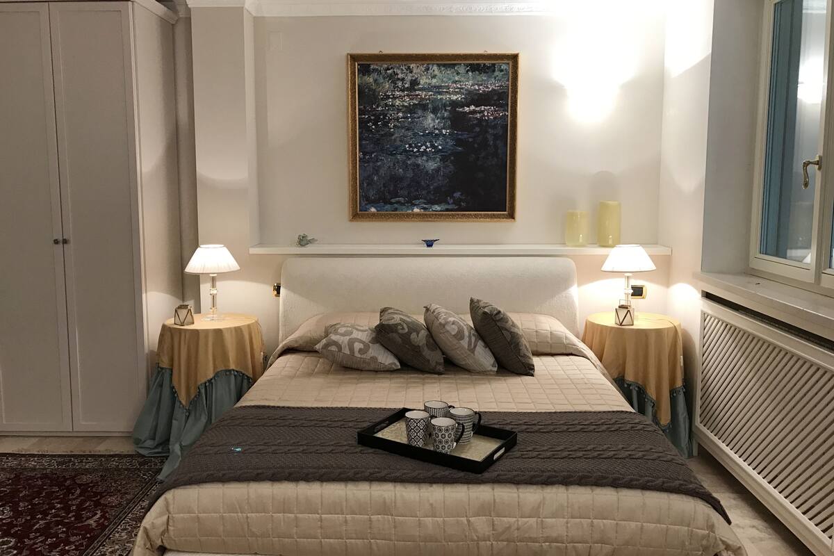 A bedroom with a large bed, two bedside tables with lamps, a framed painting above the bed, a radiator, and a wardrobe.