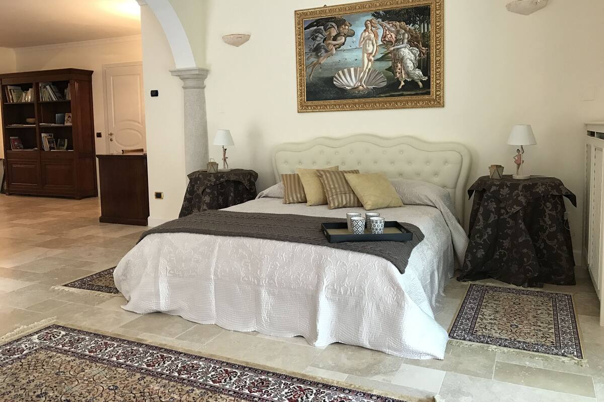 A spacious bedroom with a large bed, two nightstands, a classic painting, and a library in the background.