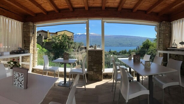 A terrace with stone walls and a view of a lake and mountains. There are white tables and chairs, and a stone fireplace. - La cascata negli ulivi (Toscolano Maderno)