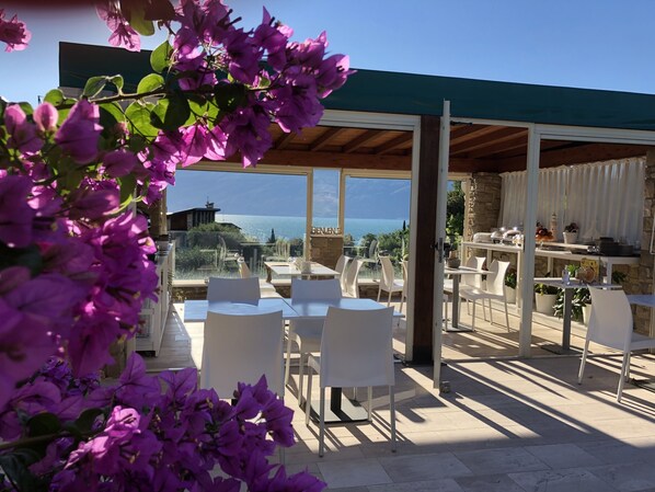 An outdoor seating area with white chairs and tables, overlooking a scenic view of the sea and mountains. - La cascata negli ulivi (Toscolano Maderno)
