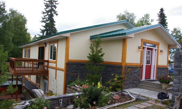 A two-story house with a stone foundation, a wooden deck, and a small garden with various plants and flowers. - Kenai Riverfront In Downtown Soldotna (Soldotna)