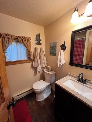 Ein Badezimmer mit Toilette, Waschbecken und Fenster mit Vorhängen. Handtücher hängen an der Wand und ein Schild mit Bergmotiv.