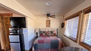 Una camera compatta arredata con un letto, un tavolino, una ventola a soffitto, una finestra con persiane, un forno a microonde, un frigorifero e una televisione su un mobile.
