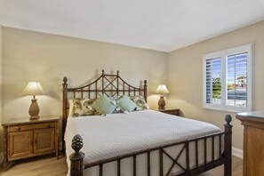 Un dormitorio con una cama de madera, dos mesitas de noche, dos lámparas, una ventana con persianas y vistas al exterior.