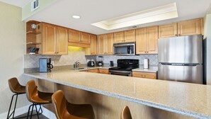 Una cocina moderna con armarios de madera, un refrigerador de acero inoxidable, un microondas y un horno empotrado. Hay tres taburetes de bar y una encimera de granito.