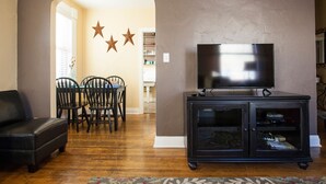 A living room with a TV, black entertainment unit, and a dining area with chairs and a table. - Charming & Historic Babs Condo on Hip Strip- Missoula's Best! (Missoula)