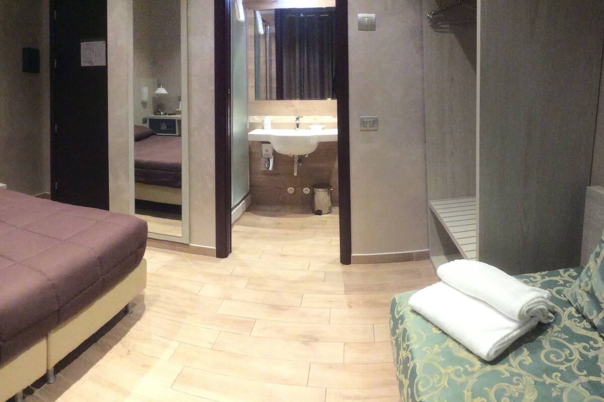 Habitación de hotel con cama, mesita de noche, silla, pequeño escritorio, espejo, lavamanos y toalla.