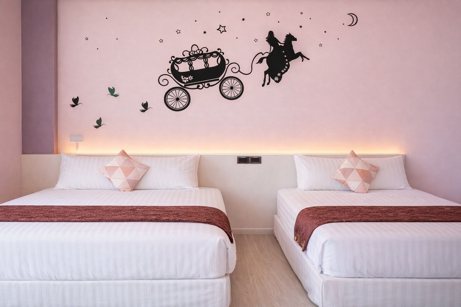 A hotel room with two beds, a nightstand, a wall decal of a horse-drawn carriage and a rider, and a wall-mounted light.