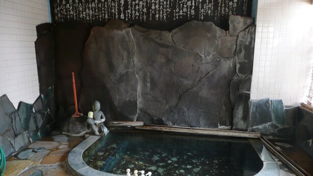 Un espace autour d’une piscine dallé de pierres, avec un grand mur de roches, une statue et une rangée de produits de toilette.