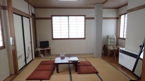 Une pièce de style japonais traditionnel, avec un sol recouvert de tatamis, une petite table basse entourée de coussins, un téléviseur et une grande fenêtre.