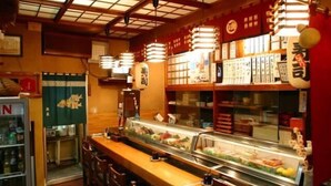 Un bar à sushis avec une vitrine réfrigérée où sont exposés différents types de fruits de mer, des tables en bois et des lanternes en papier suspendues.