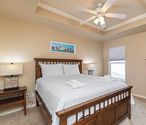 A bedroom with a large bed, a ceiling fan, a painting, a TV, and a wooden dresser.