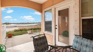 A covered porch with two patterned chairs, a door with a screen, and a view of a road and beach. - Spacious Family Retreat | Pool Access & Beach Nearby (Corpus Christi)