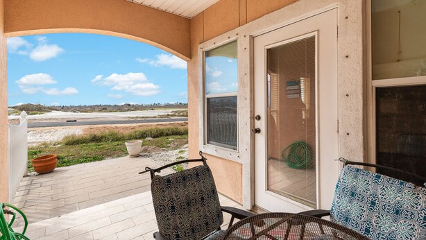 A covered porch with two patterned chairs, a door with a screen, and a view of a road and beach. - Spacious Family Retreat | Pool Access & Beach Nearby (Corpus Christi)