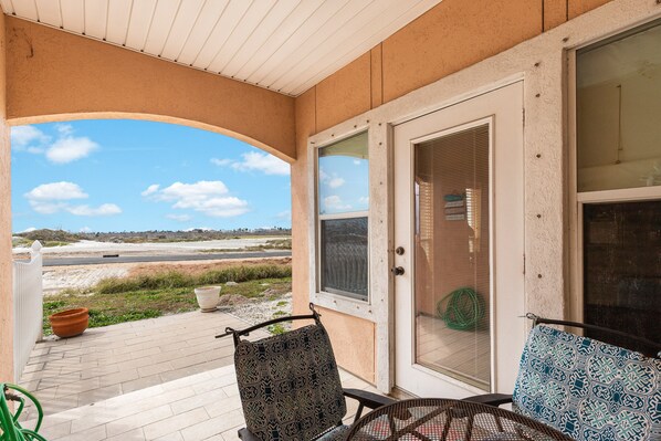 Eine überdachte Veranda mit zwei gemusterten Stühlen, einer Glastür und Blick auf eine Straße und den Strand.