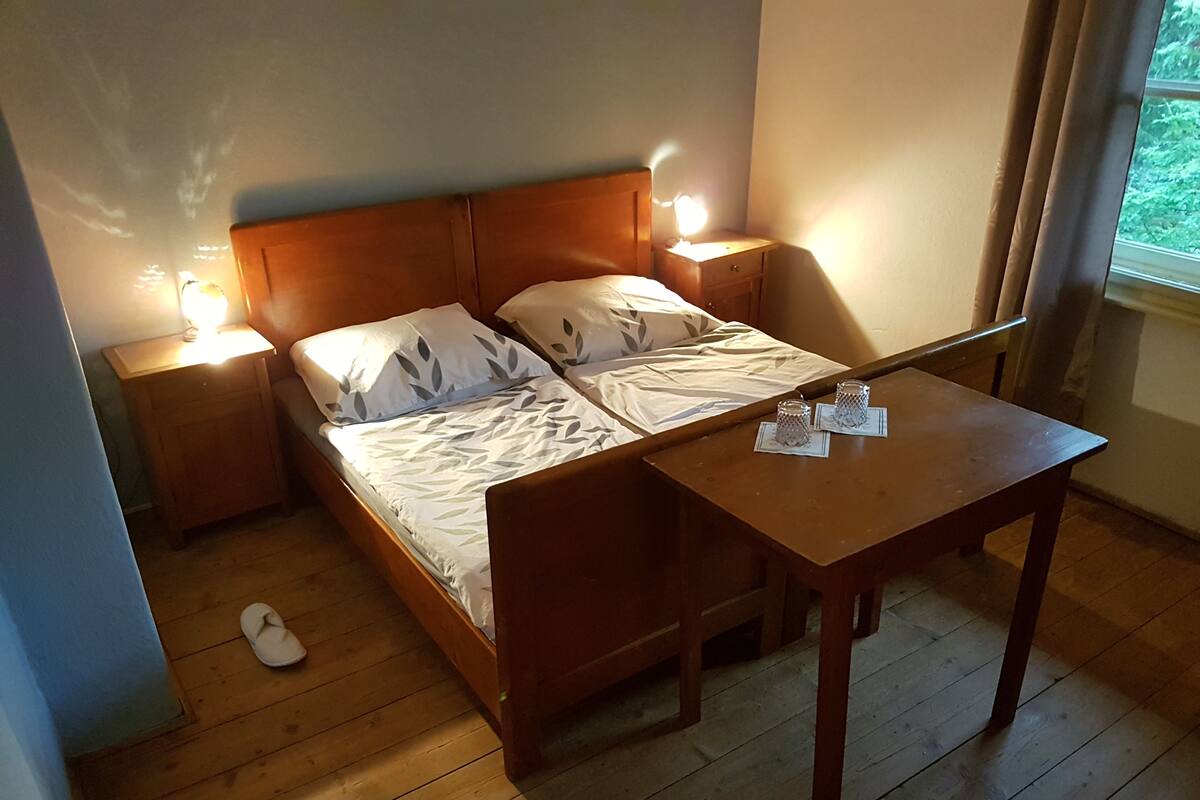 A wooden bed with a leaf-patterned bedspread, two bedside tables with lamps, and a small wooden table with two glasses.