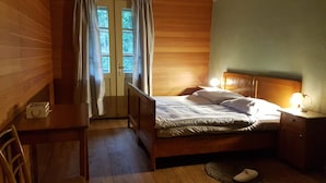 A small, cozy bedroom with a wooden bed, a nightstand with a lamp, a desk, and a chair. There is a window with curtains and a door leading outside. - Vila Koman (Jezersko)