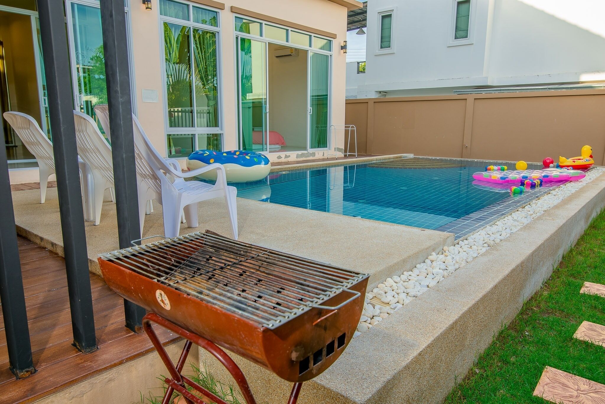 A swimming pool with lounge chairs and inflatable toys, a barbecue grill, and a house with large windows.
