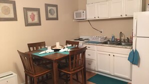 Una cocina pequeña con armarios blancos, un microondas, una estufa y un refrigerador. Hay una mesa de comedor con sillas y un mantel individual azul.