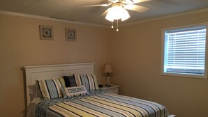 Un dormitorio con una cama, un ventilador de techo, una ventana con persianas y dos cuadros colgados en la pared.