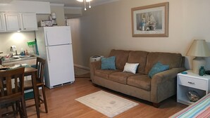 Un espacio habitable compacto con kitchenette, un sofá marrón con cojines azules, una mesa de comedor con sillas y un ventilador de techo.