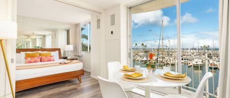 A modern hotel room with a large balcony, a bed, a dining table with chairs, and a view of a marina.