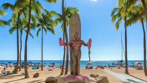 Una estatua de un hombre sosteniendo una tabla de surf en una playa con palmeras y un cielo despejado.