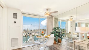 Una habitación amplia con un gran ventanal que da a un puerto deportivo, ventilador de techo y comedor con sillas blancas y una mesa.